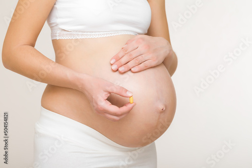 Young adult pregnant woman hand holding pill for future newborn health on big naked belly. Receiving vitamins in pregnancy time. Baby expectation. Closeup. Isolated on light gray background.