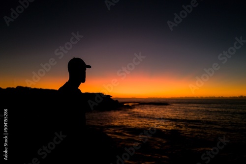 Sillhouette of man wearing baseball cap looking out to the ocean at sunrise
