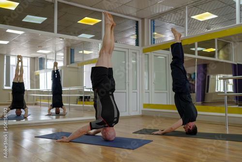 Two men stand inverted on the top of their head, doing Shirshasana, also known as headstand.