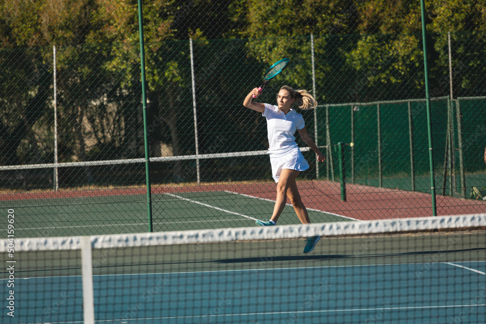 Young female caucasian player jumping while playing tennis game at court on sunny day