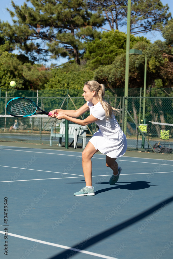Confident young female caucasian tennis player hitting ball with racket at court on sunny day