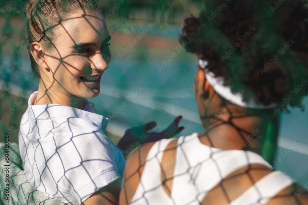 Smiling caucasian female player talking with african american athlete at tennis court during break