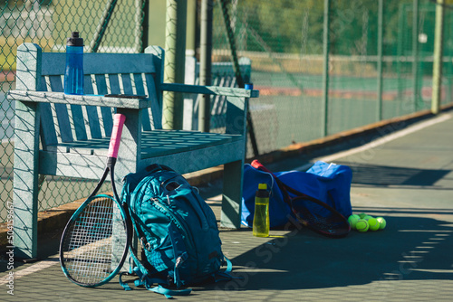 Tennis racket with backpacks and bottles by empty wooden bench at court on sunny day