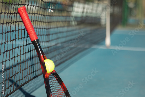 Tennis ball and red racket leaning on net at court on sunny day