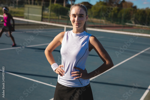 Portrait of smiling confident young female player standing with hands on hips at tennis court