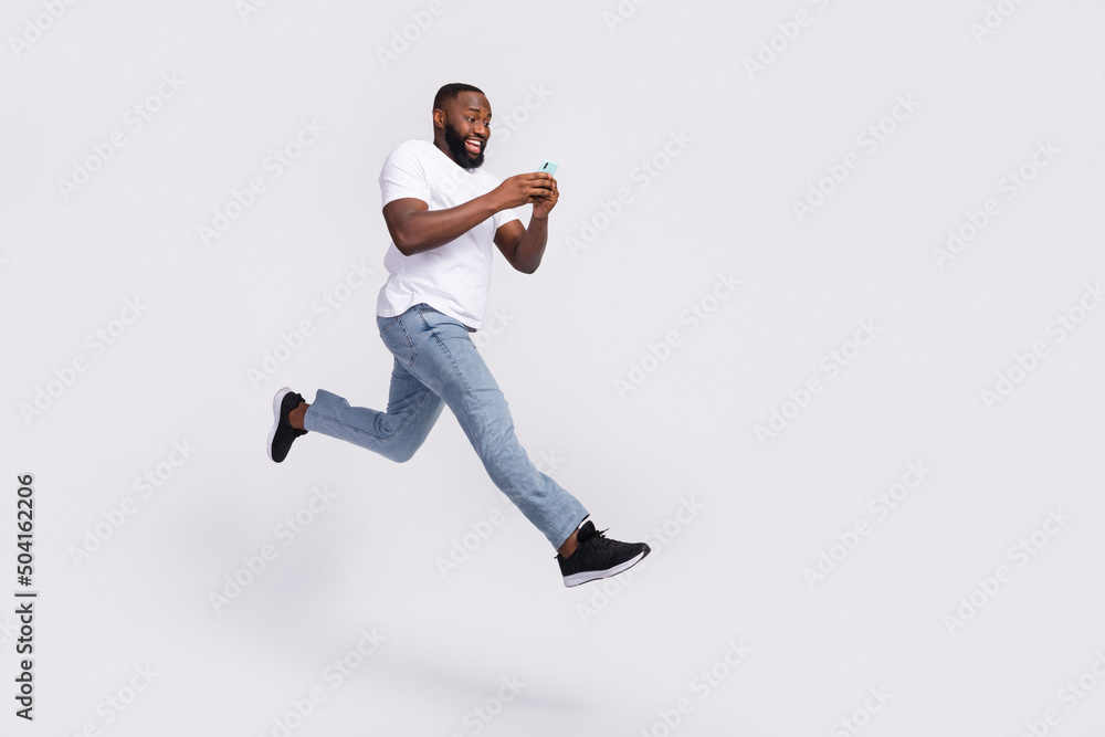 Photo of funky charming guy dressed t-shirt jumping high chatting modern gadget empty space isolated white color background Photo of funky charming guy dressed t-shirt jumping high chatting modern gadget empty space isolated white color background