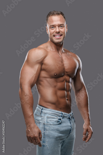 Studio shot of handsome guy with muscular build dressed in jeans posing against grey background.