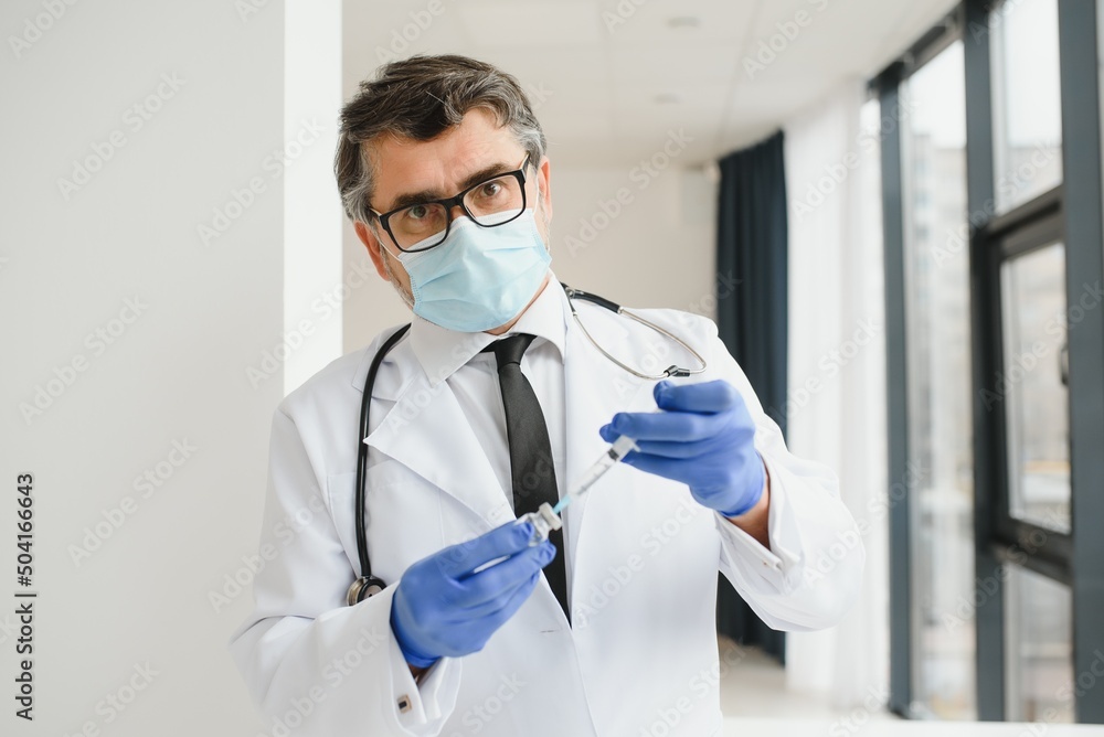Mid aged male doctor wearing protective mask holding a syringe with vaccine. Mid aged male doctor wearing protective mask holding a syringe with vaccine.