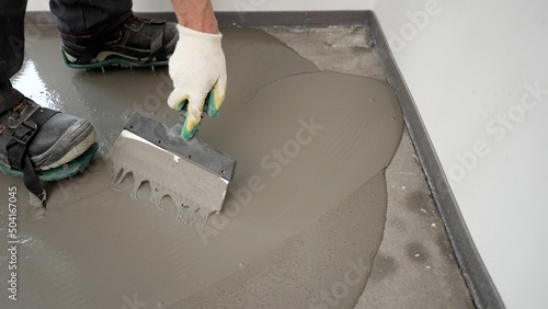 Repairman in protective gloves using a metal spatula to level a liquid self-levelling floor screed. A worker pours liquid cement mortar at a construction site.
