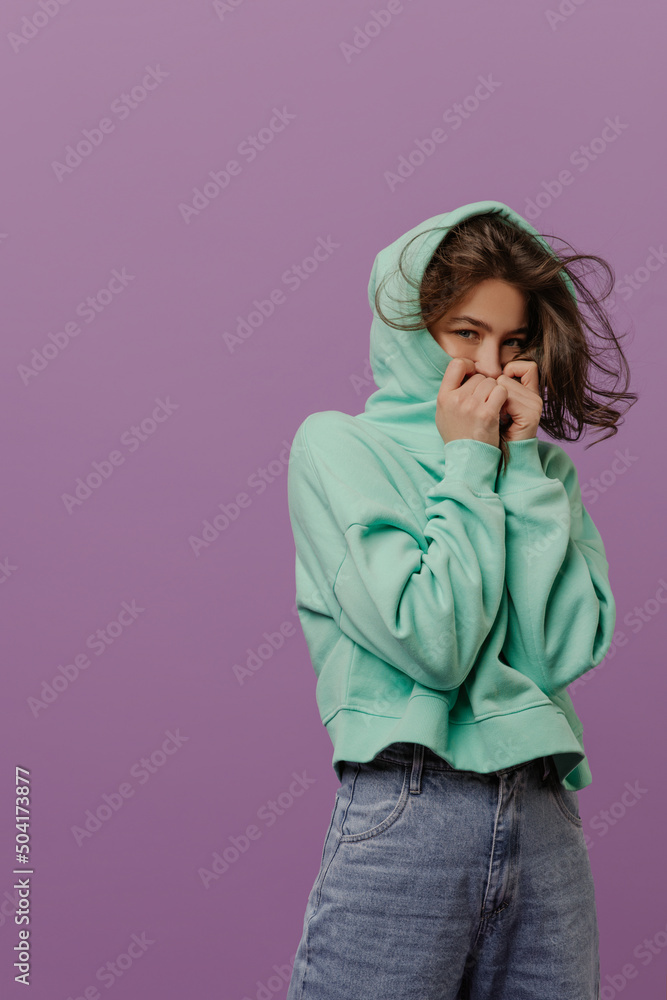 European young girl with dark hair hides face covered with hood looking at camera in studio. Brunette wears casual clothes of hoodies and jeans. Lifestyle concept European young girl with dark hair hides face covered with hood looking at camera in studio. Brunette wears casual clothes of hoodies and jeans. Lifestyle concept