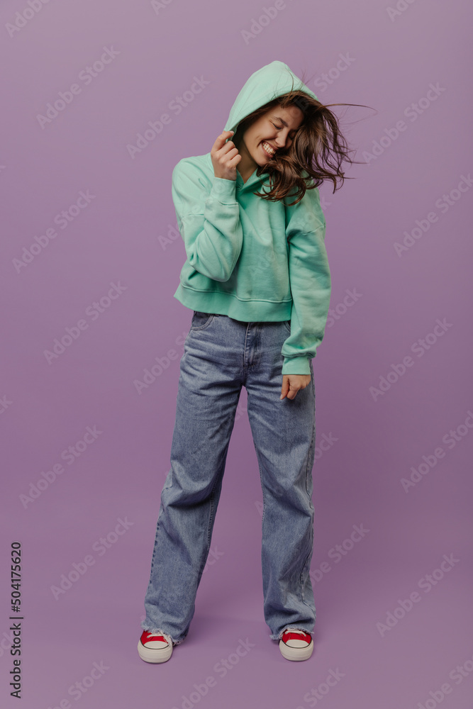 Full-length, funny young caucasian girl laughs with eyes closed and hood over head standing in studio. Brunette girl with disheveled hair wear hoodie and jeans. People's emotions, lifestyle concept. Full-length, funny young caucasian girl laughs with eyes closed and hood over head standing in studio. Brunette girl with disheveled hair wear hoodie and jeans. People's emotions, lifestyle concept.