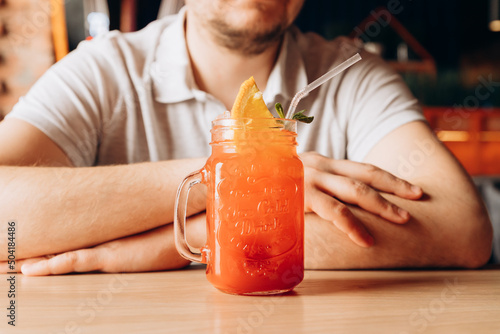 Young caucasian man and Red Strawberry lemonade with ice,  cocktail tube and orange slice in glass with handle on wooden table. Male hands and cocktail. Front view, lifestyle