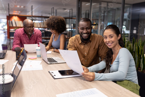 Portrait of smiling biracial business colleagues sitting with document by professionals at office