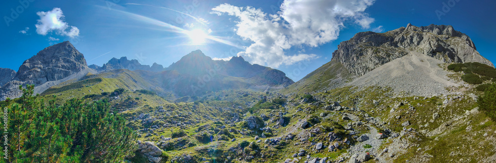 Obraz premium Ein weites Panorama zeigt eine beeindruckende Gebirgskette unter strahlend blauem Himmel, während die Sonne hell über den schroffen, teils begrünten Felsgipfeln und einem steinigen Tal leuchtet.