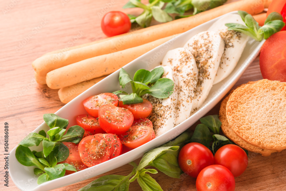 Traditional italian caprese salad with baby tomatoes and sliced mozzarella, basil, olive oil, corn salad, bruschettas and grissini on wooden table background. Classic restaurant appetizer Traditional italian caprese salad with baby tomatoes and sliced mozzarella, basil, olive oil, corn salad, bruschettas and grissini on wooden table background. Classic restaurant appetizer