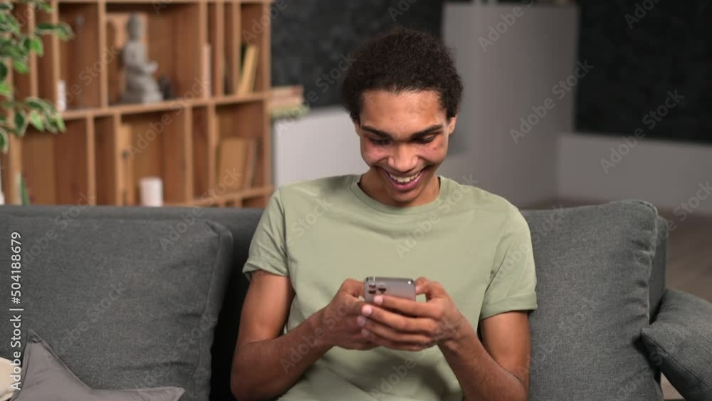 Handsome man sending messages on the smartphone, sitting on the comfortable couch with a cell phone in hands, multiracial teenager guy texting, scrolling and web surfing Handsome man sending messages on the smartphone, sitting on the comfortable couch with a cell phone in hands, multiracial teenager guy texting, scrolling and web surfing