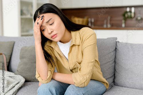 Stressed young asian woman touching head and feels dizzy, suffering from headache and migraine pain, sits on the sofa at home, tired and exhausted lady feels bad