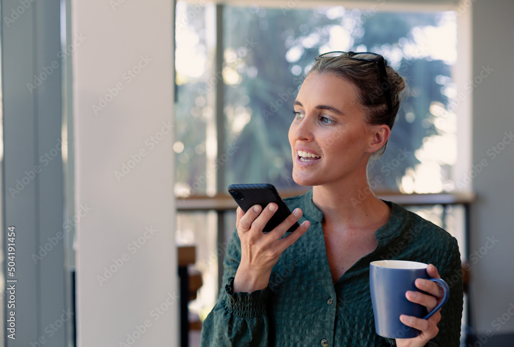 Caucasian young female advisor holding coffee mug talking through smartphone speaker at workplace Caucasian young female advisor holding coffee mug talking through smartphone speaker at workplace