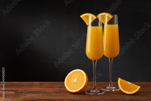 A pair of mimosa cocktail in flute glass with orange slice on a wooden bar and dark background.