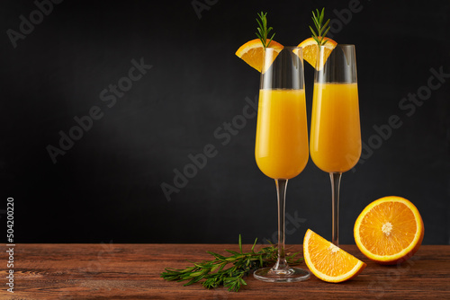 A pair of mimosa cocktail in flute glass with orange slice and rosemary twig on a wooden bar and dark background.