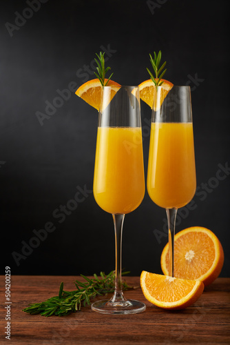 A pair of mimosa cocktail in flute glass with orange slice and rosemary twig on a wooden bar and dark background.