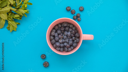 Pink cup full of blueberries on a turquoise blue background and with mint leaves in the upper left corner
