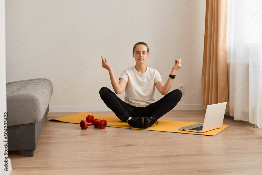 Concentrated Caucasian woman sit on gymnastic mat in lotus pose near laptop, meditate and listen to calm music, young girl practice yoga at home, relaxing calming down. Concentrated Caucasian woman sit on gymnastic mat in lotus pose near laptop, meditate and listen to calm music, young girl practice yoga at home, relaxing calming down.