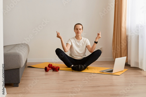 Concentrated Caucasian woman sit on gymnastic mat in lotus pose near laptop, meditate and listen to calm music, young girl practice yoga at home, relaxing calming down.