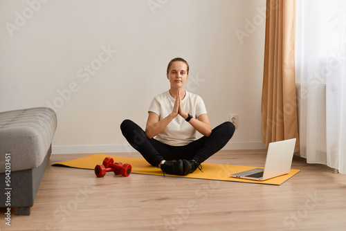 Calm relaxed woman sit on gymnastic mat on floor with crossed legs using laptop for listen to calm music or watching tutorial video, keeps palms together in praying gesture.