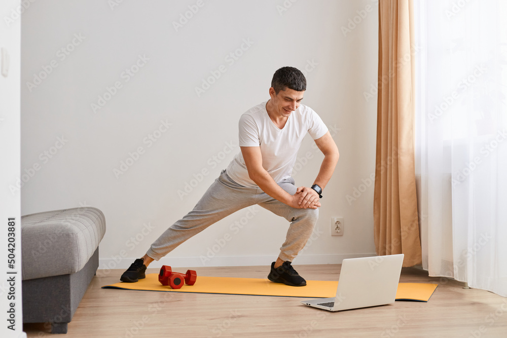 Fit gymnast man with dark hair in sportswear standing on mat and stretching legs, warming up, doing sports flexibility exercises, fitness and yoga workout at home. Fit gymnast man with dark hair in sportswear standing on mat and stretching legs, warming up, doing sports flexibility exercises, fitness and yoga workout at home.