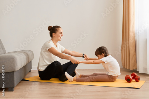 Woman with little daughter wearing sportswear doing sport exercises at home, holding hands together and practicing yoga at home, doing stretching exercise together. Healthy lifestyle.