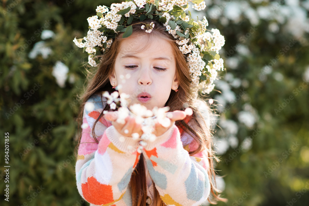 Happy smiling kid girl 6-7 year old wear knit colorful sweater and floral wreath hairstyle with flowers in park over nature background. Springtime. Spring season. Childhood. Child blow on petals