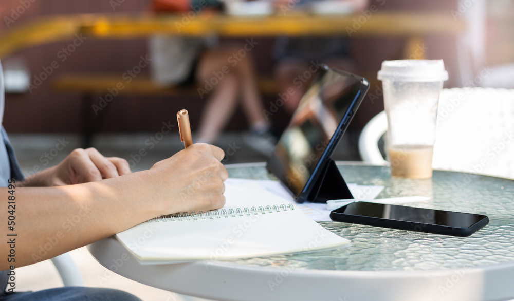 Close up view, hand of asian woman writing her plan on notebook and using digital tablet working online with paperwork of investment on glass table