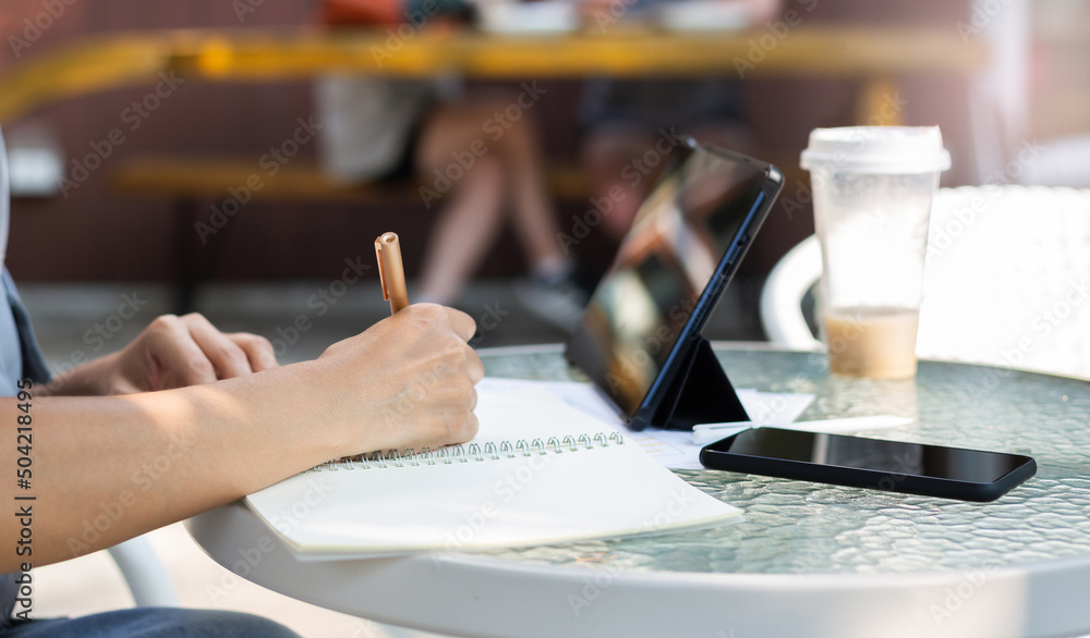 Close up view, hand of asian woman writing her plan on notebook and using digital tablet working online with paperwork of investment on glass table Close up view, hand of asian woman writing her plan on notebook and using digital tablet working online with paperwork of investment on glass table