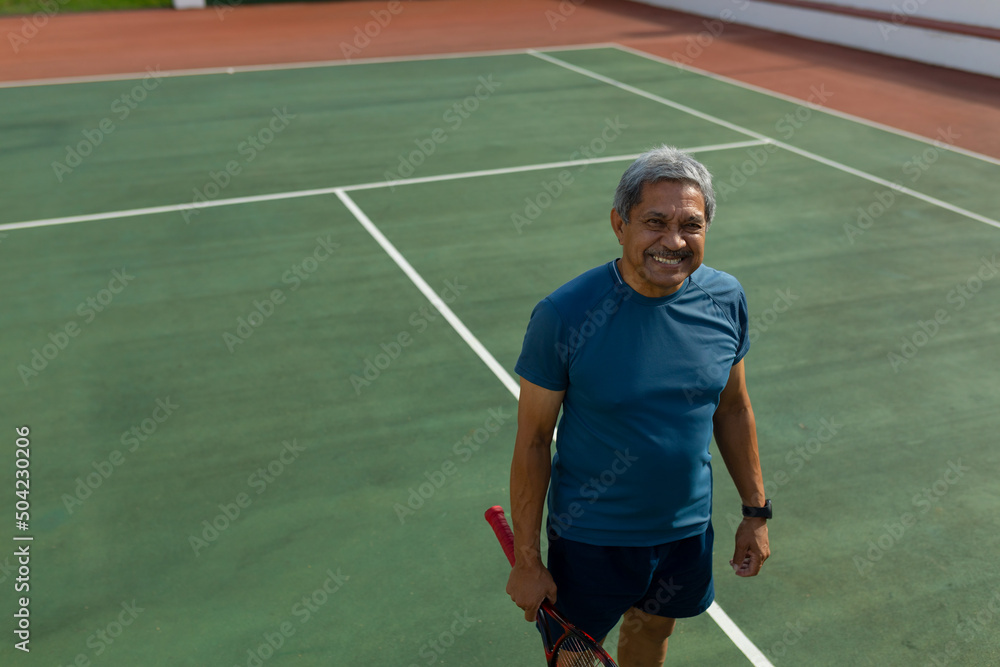 High angle portrait of smiling biracial senior man with racket standing in tennis court on sunny day