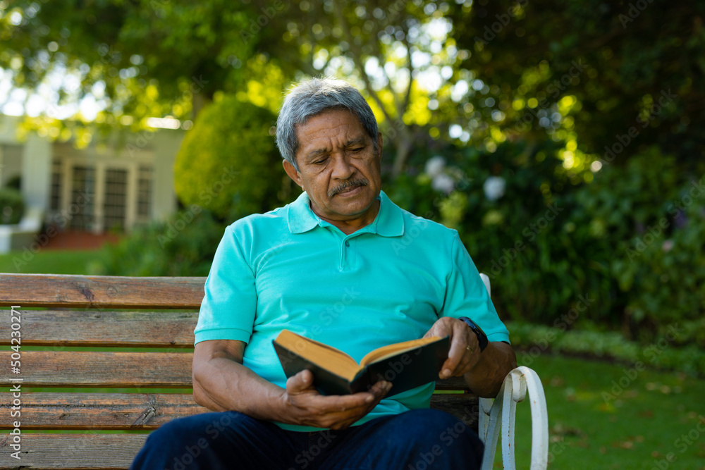 Obraz premium Serious biracial senior man reading book with focus while sitting on bench against plants in park