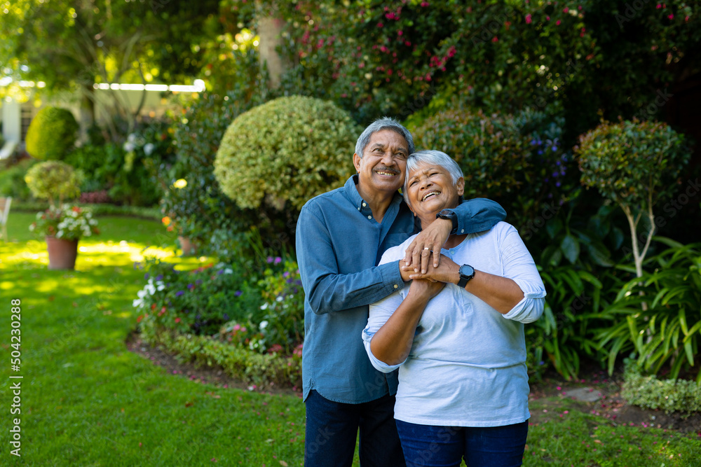 Fototapeta premium Smiling biracial senior couple embracing and looking up while standing against plants in park