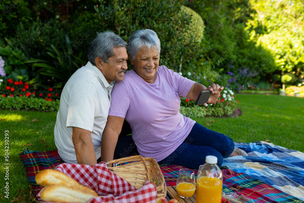 Obraz premium Biracial senior woman taking selfie with husband over mobile phone while sitting on blanket in park