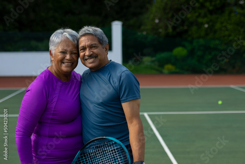 Portrait of smiling biracial senior couple embracing while standing in tennis court