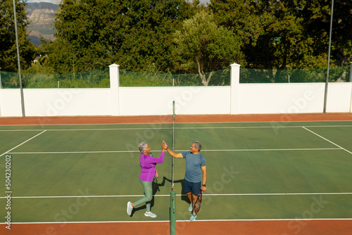 High angle view of biracial senior couple giving high-five while playing tennis in court in summer
