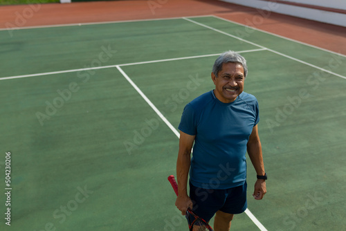 High angle portrait of smiling biracial senior man with racket standing in tennis court on sunny day © wavebreak3