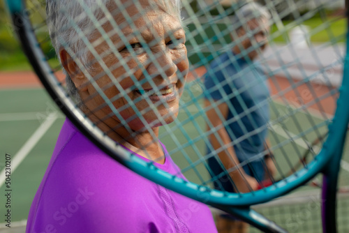 Close-up portrait of biracial smiling senior woman with man looking through tennis racket in court © wavebreak3