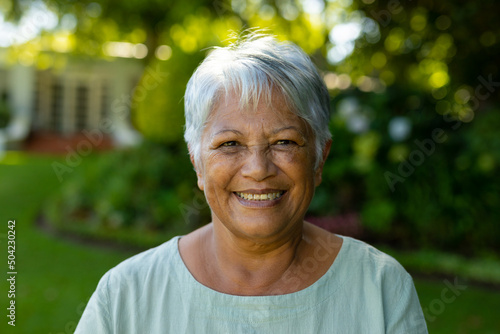 Close-up portrait of smiling biracial senior woman with short gray hair against trees in park