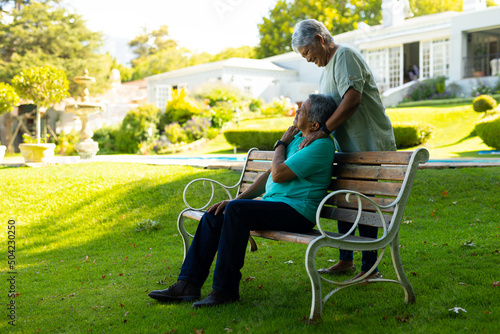 Cheerful biracial senior woman standing and looking at senior husband sitting on bench in park
