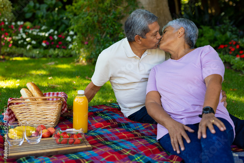 Biracial senior couple kissing while sitting with bread, fruits and juice on blanket in park