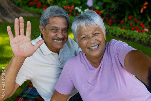 Portrait of cheerful biracial senior man waving hand while enjoying picnic with senior wife in park
