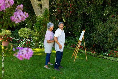 Biracial senior woman tying husband's apron standing by canvas and and easel against plants in yard
