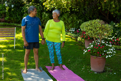 Smiling biracial senior couple holding hands while standing on yoga mats against plants in yard