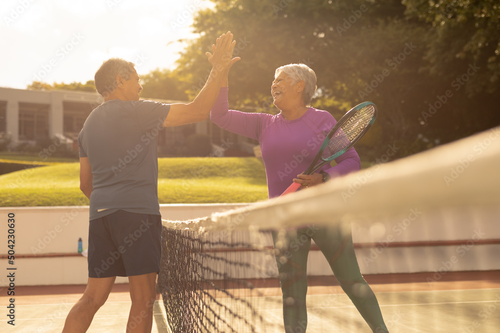 Cheerful biracial senior couple giving high-five while playing tennis at court during sunset - obrazy, fototapety, plakaty 