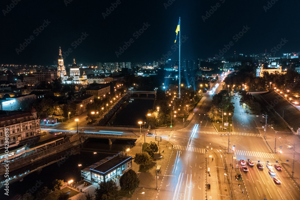 Illuminated flag of Ukraine and Holy Annunciation Cathedral on river Lopan at night. Kharkiv city aerial, Ukraine in long exposure